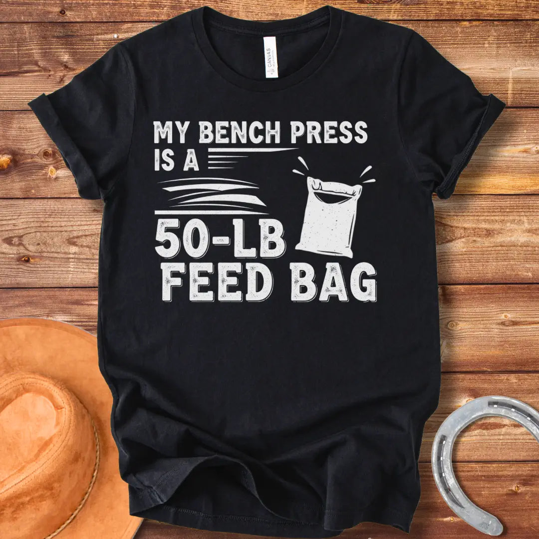 A graphic design on a t-shirt showing a humorous depiction of a feed bag labeled as a '50-lb' weight for a bench press.