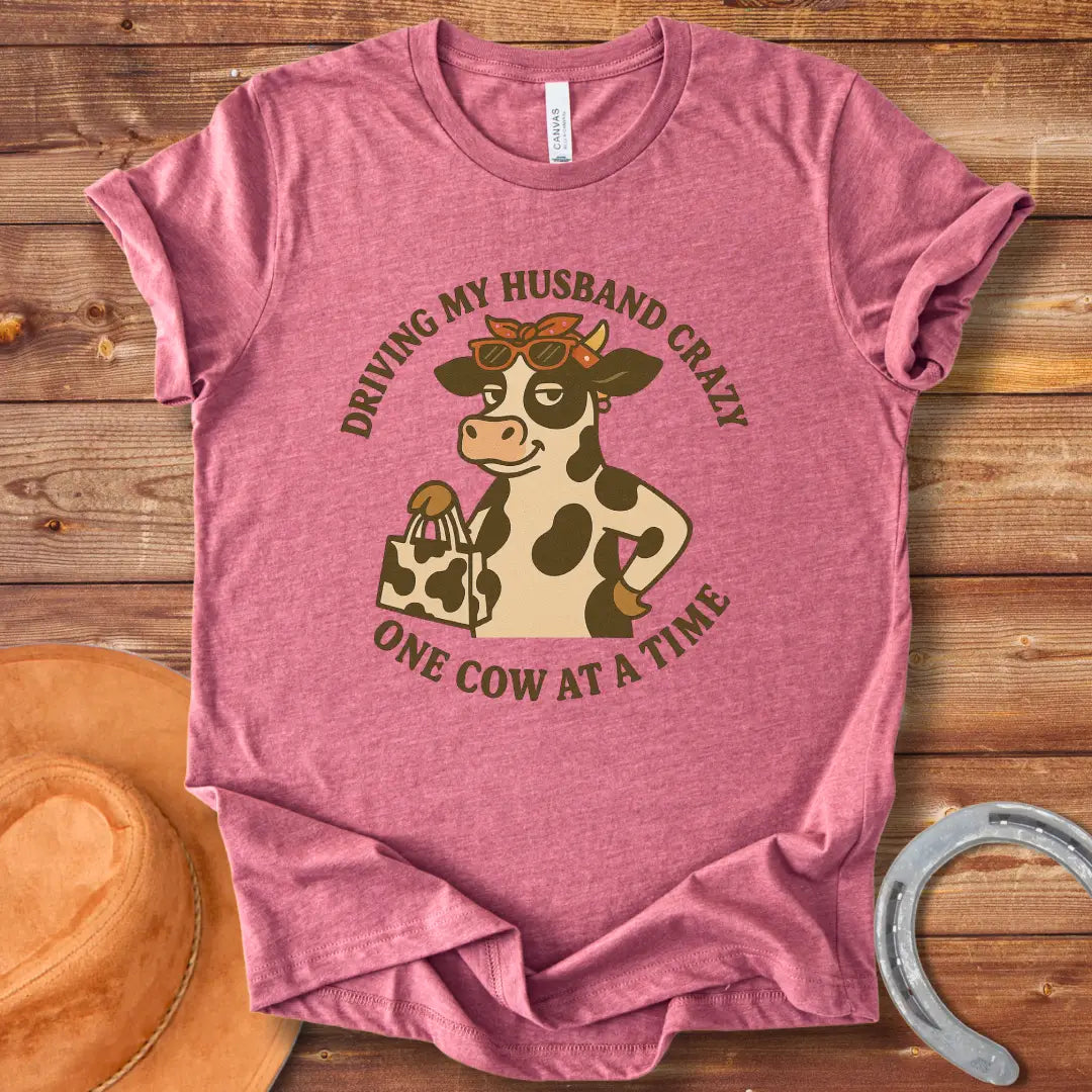 A cartoon cow wearing sunglasses and a bandana, holding a shopping bag, with the text 'Driving My Husband Crazy One Cow At A Time' around it.
