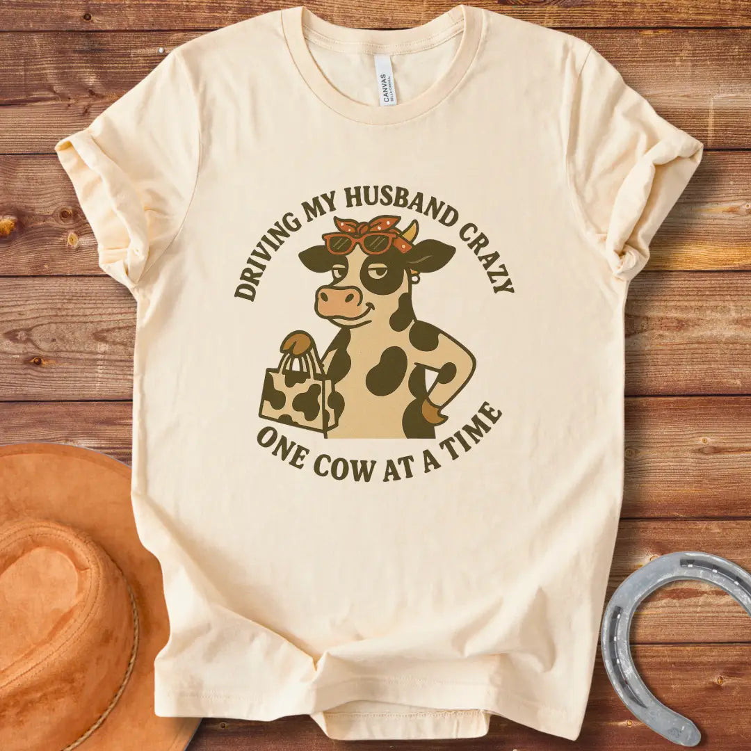 A cartoon cow wearing sunglasses and a bandana, holding a shopping bag, with the text 'Driving My Husband Crazy One Cow At A Time' around it.