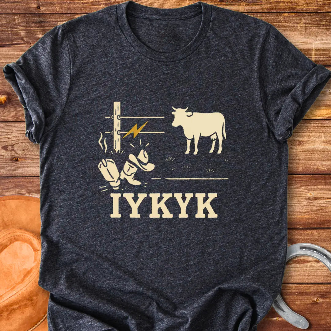 Graphic design on a t-shirt depicts a shocked cowboy hat and boots with a hot wire and a cow in the background, with the words IYKYK at the bottom