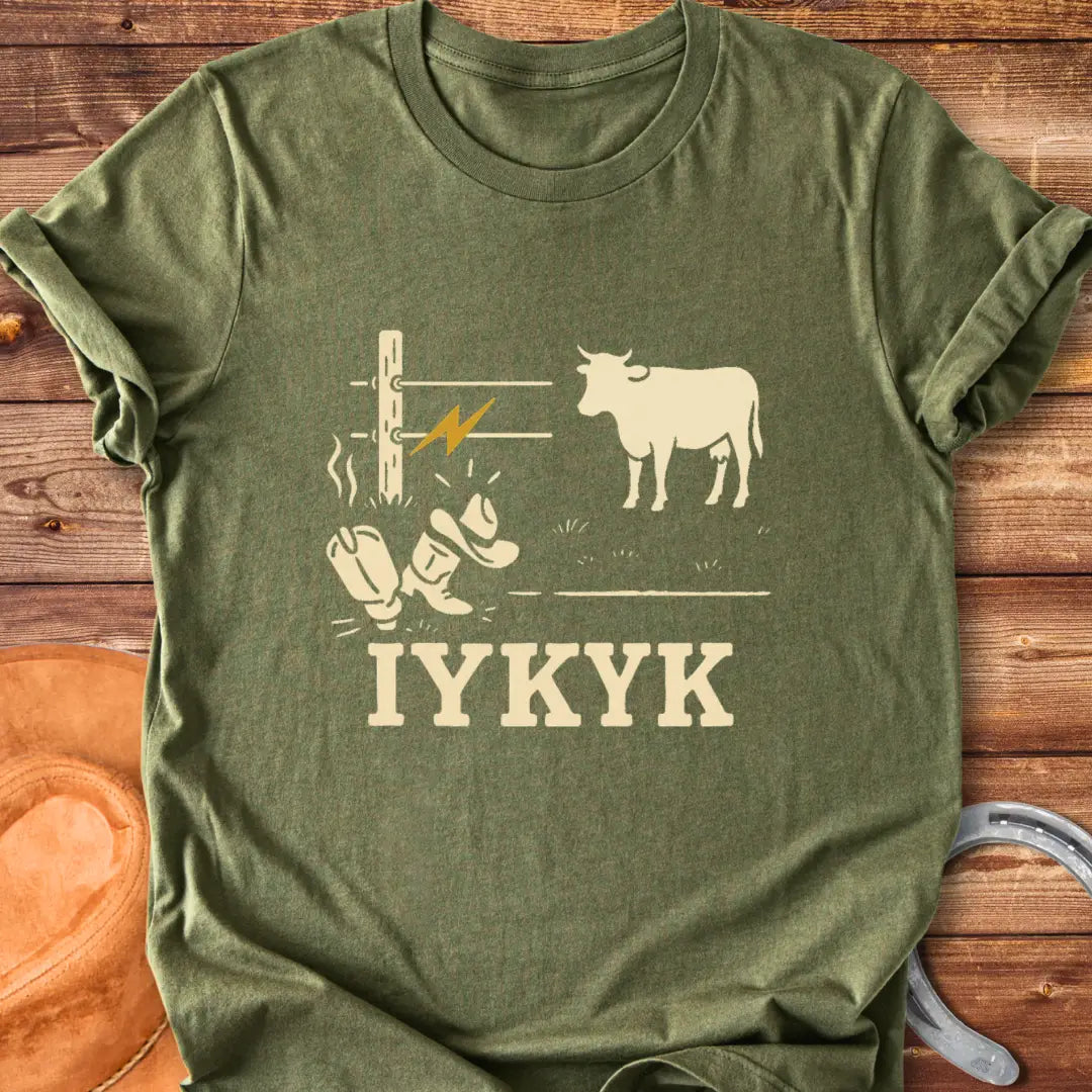 Graphic design on a t-shirt depicts a shocked cowboy hat and boots with a hot wire and a cow in the background, with the words IYKYK at the bottom