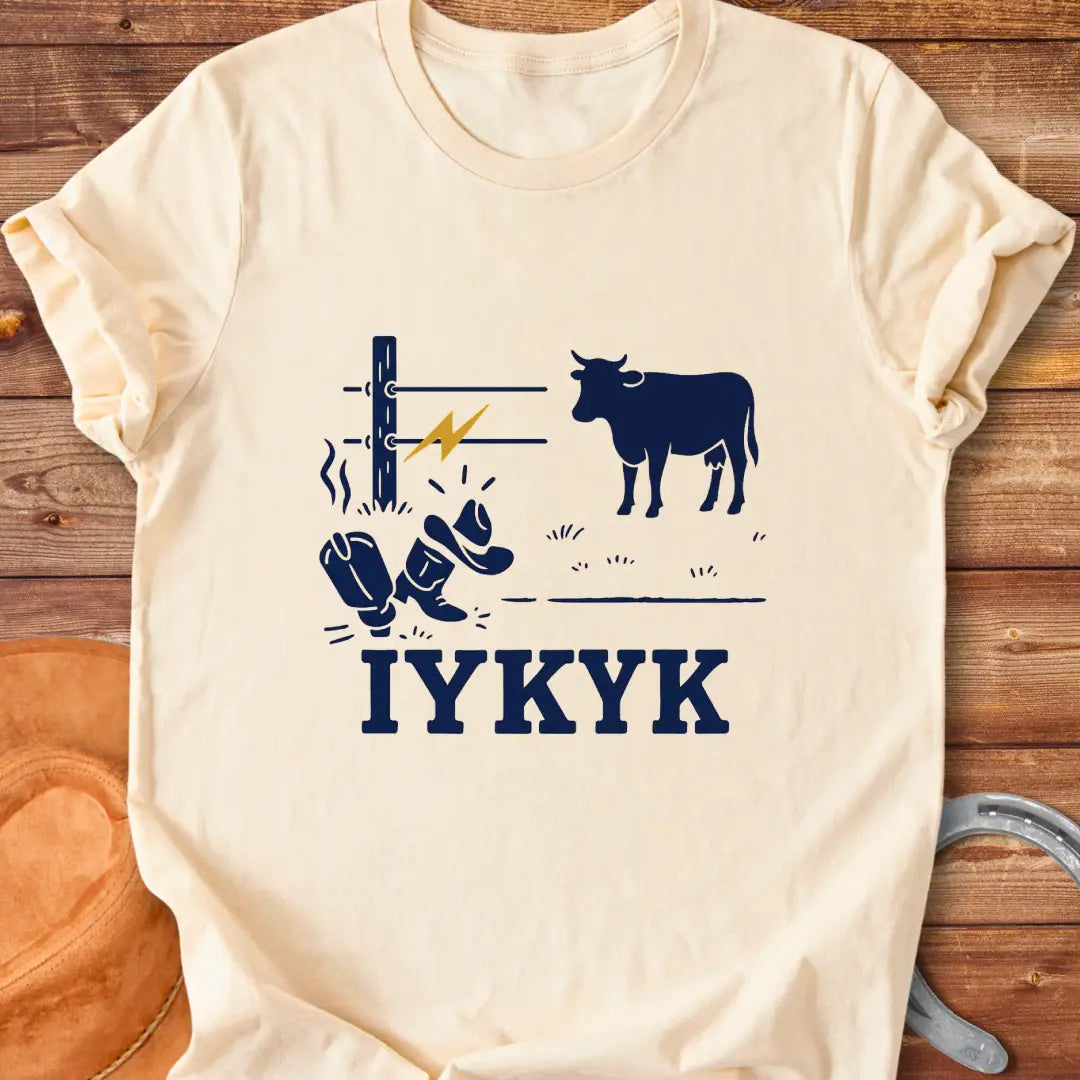 Graphic design on a t-shirt depicts a shocked cowboy hat and boots with a hot wire and a cow in the background, with the words IYKYK at the bottom
