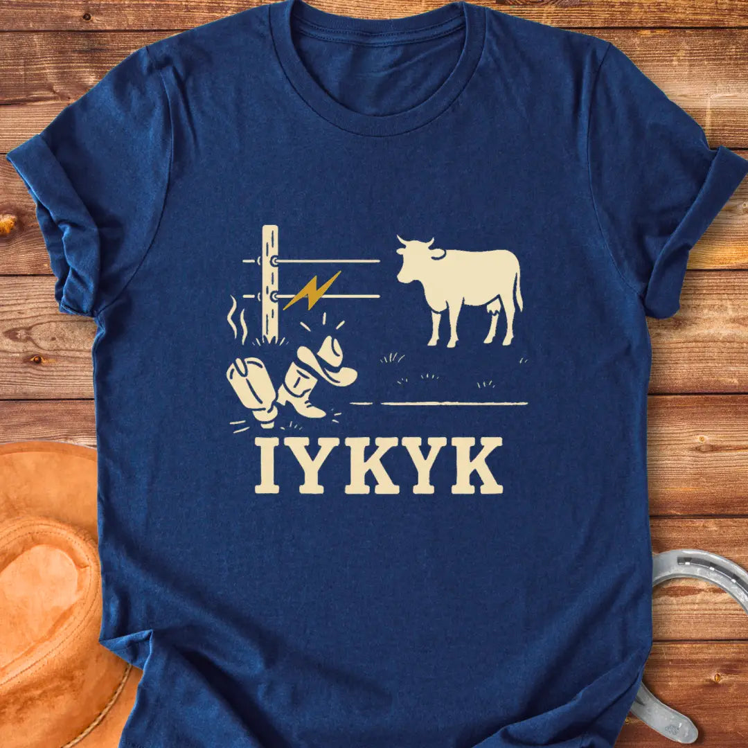 Graphic design on a t-shirt depicts a shocked cowboy hat and boots with a hot wire and a cow in the background, with the words IYKYK at the bottom
