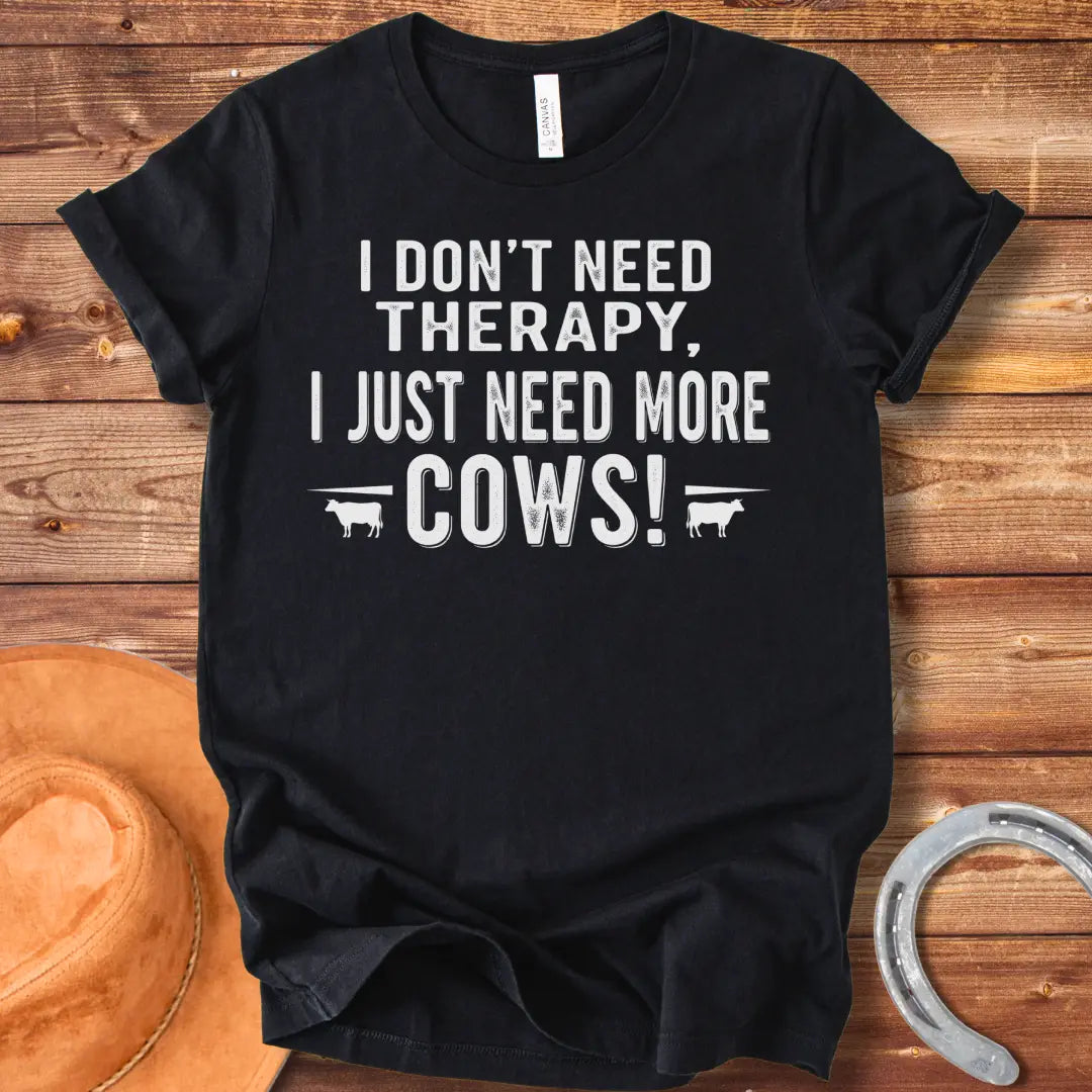 A black and white graphic tee design that says 'I DON'T NEED THERAPY, I JUST NEED MORE COWS!' with two small cow illustrations flanking the text.
