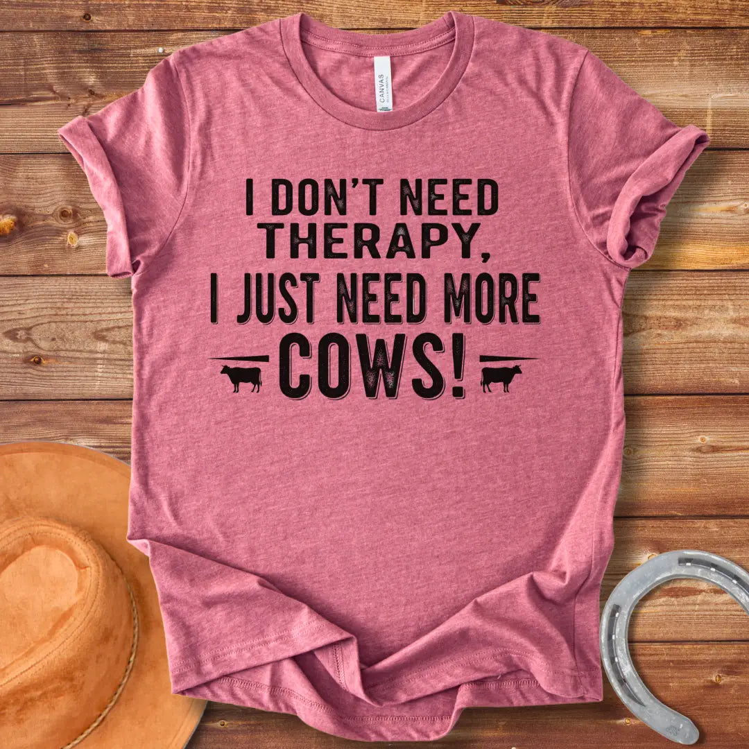 A black and white graphic tee design that says 'I DON'T NEED THERAPY, I JUST NEED MORE COWS!' with two small cow illustrations flanking the text.
