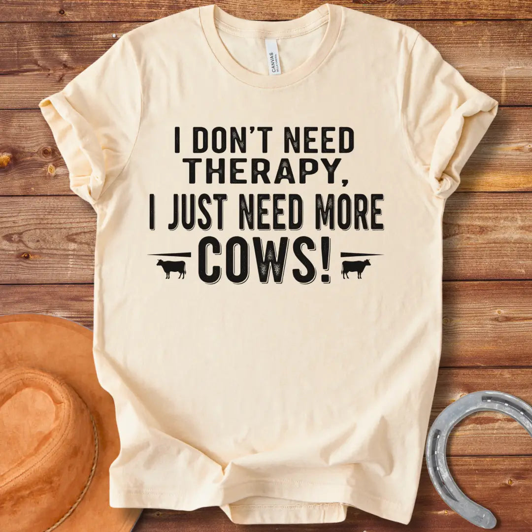 A black and white graphic tee design that says 'I DON'T NEED THERAPY, I JUST NEED MORE COWS!' with two small cow illustrations flanking the text.