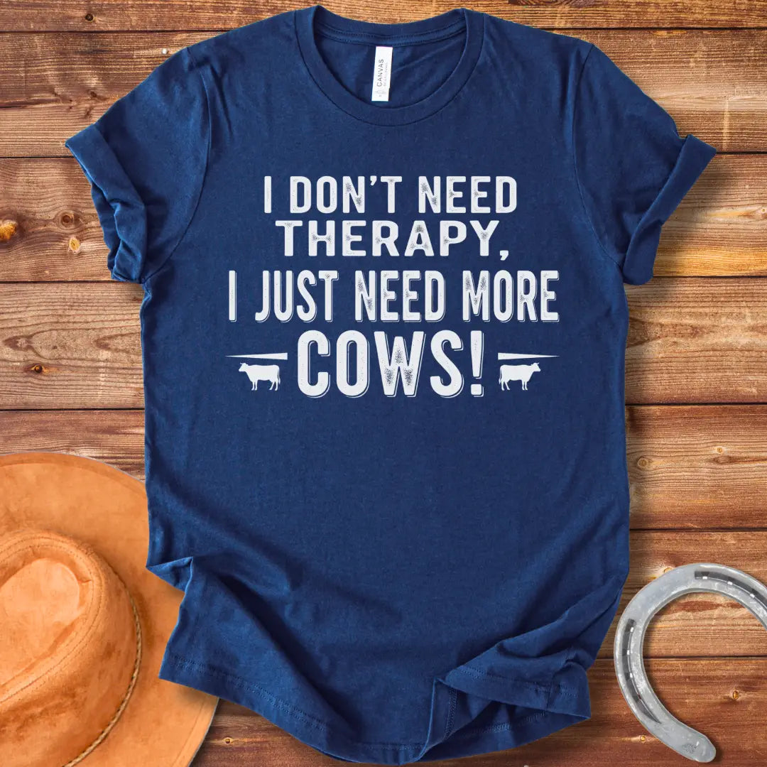A black and white graphic tee design that says 'I DON'T NEED THERAPY, I JUST NEED MORE COWS!' with two small cow illustrations flanking the text.