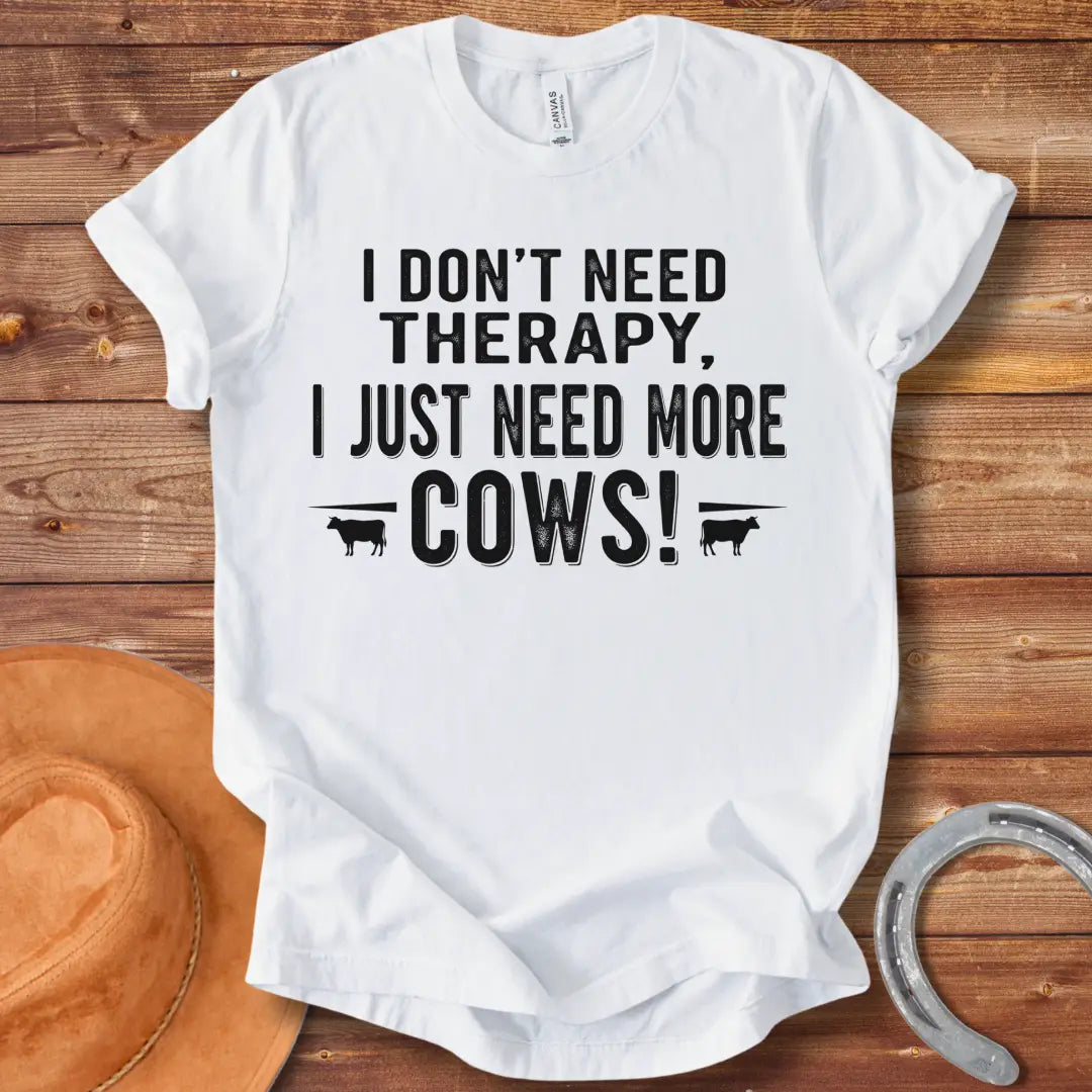 A black and white graphic tee design that says 'I DON'T NEED THERAPY, I JUST NEED MORE COWS!' with two small cow illustrations flanking the text.
