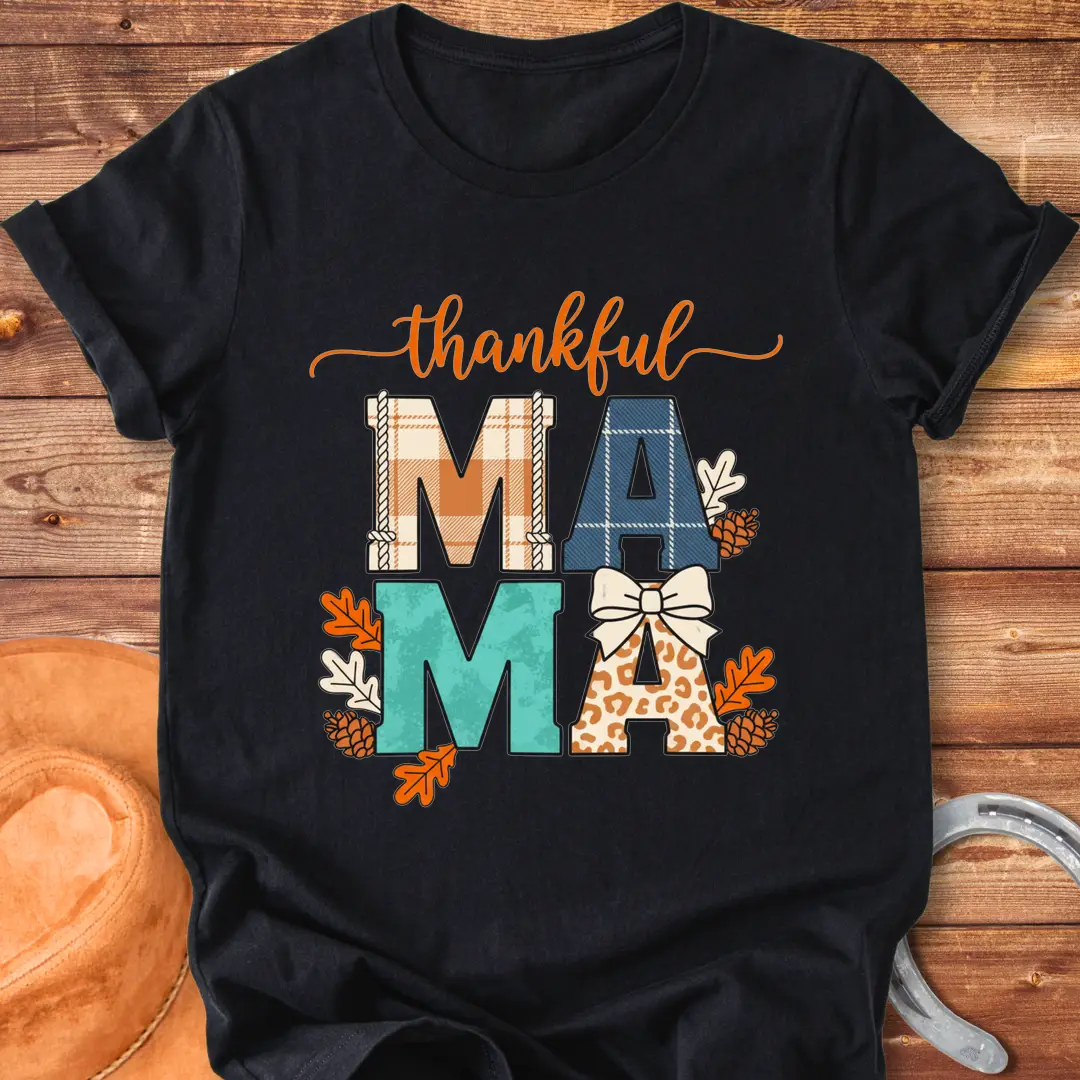 Patchwork Thankful Mama T-Shirt with autumn leaves, pinecones, and a bow on a black background. The design features the word 'thankful' and 'MAMA' in plaid and animal print letters.