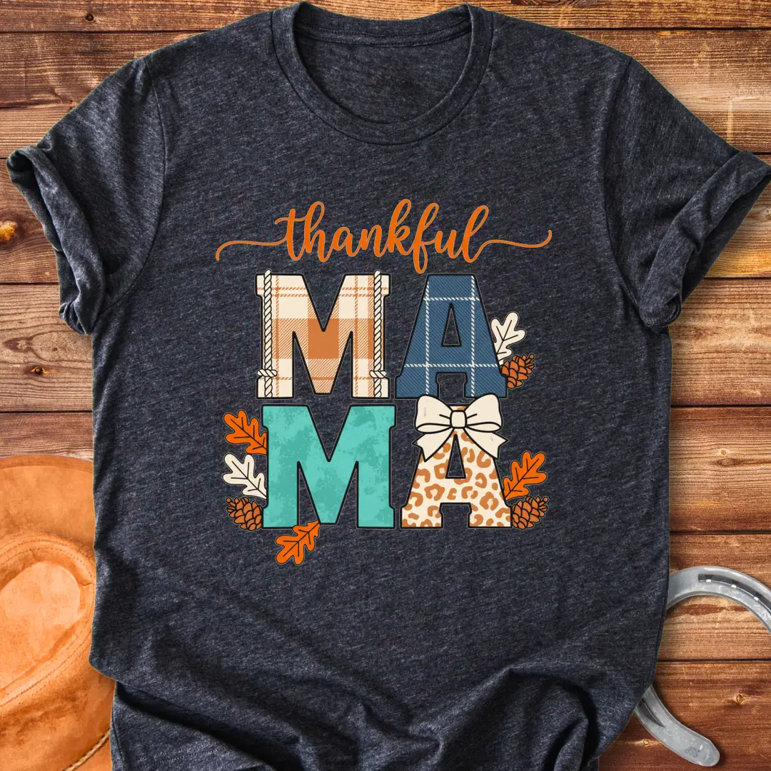 Patchwork Thankful Mama T-Shirt with autumn leaves, pinecones, and a bow on a black background. The design features the word 'thankful' and 'MAMA' in plaid and animal print letters.