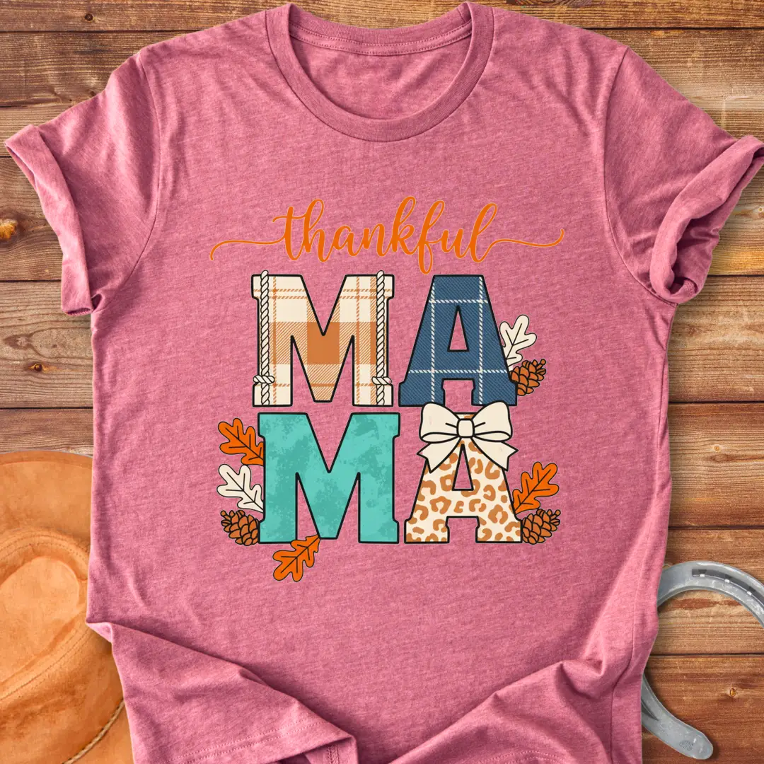 Patchwork Thankful Mama T-Shirt with autumn leaves, pinecones, and a bow on a black background. The design features the word 'thankful' and 'MAMA' in plaid and animal print letters.