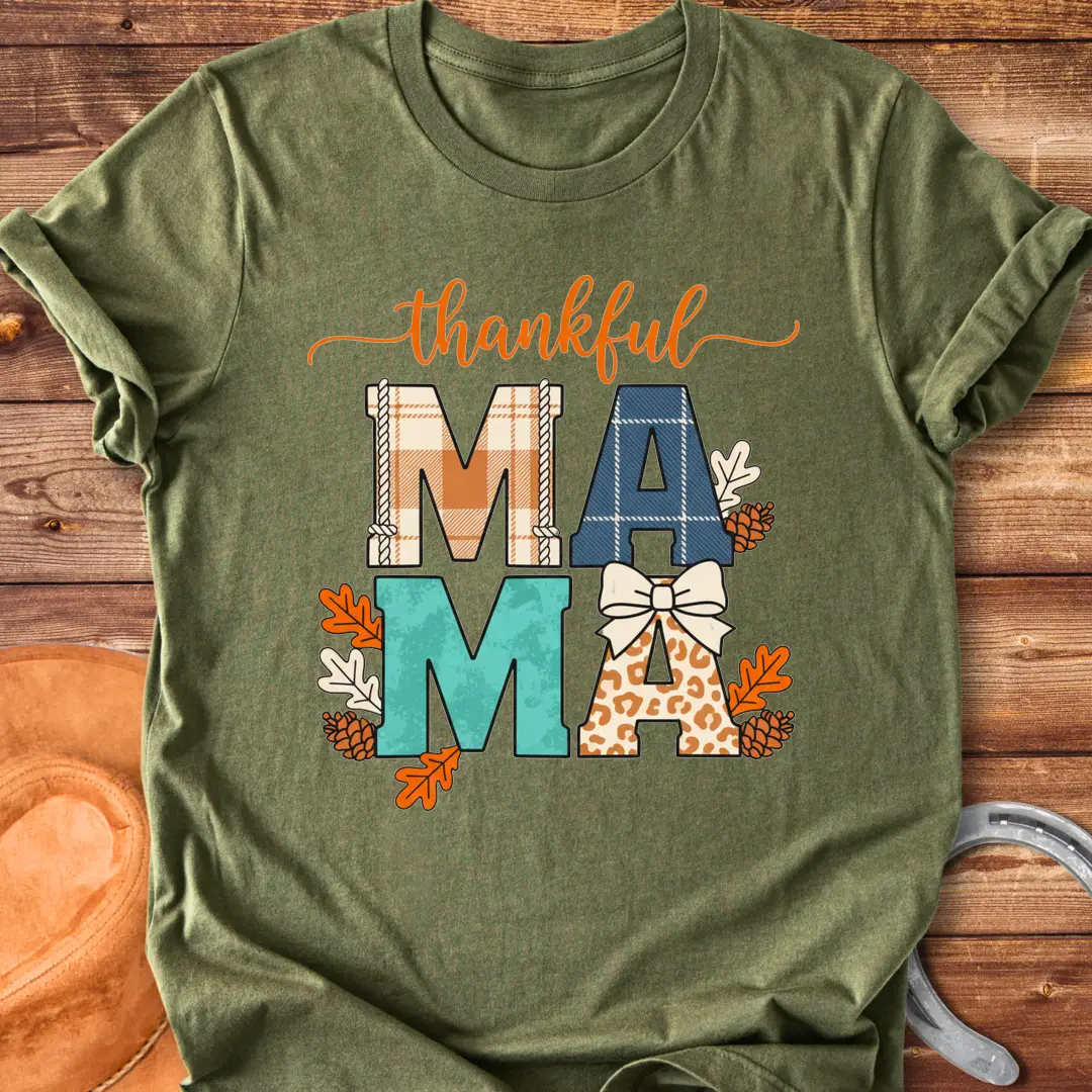 Patchwork Thankful Mama T-Shirt with autumn leaves, pinecones, and a bow on a black background. The design features the word 'thankful' and 'MAMA' in plaid and animal print letters.
