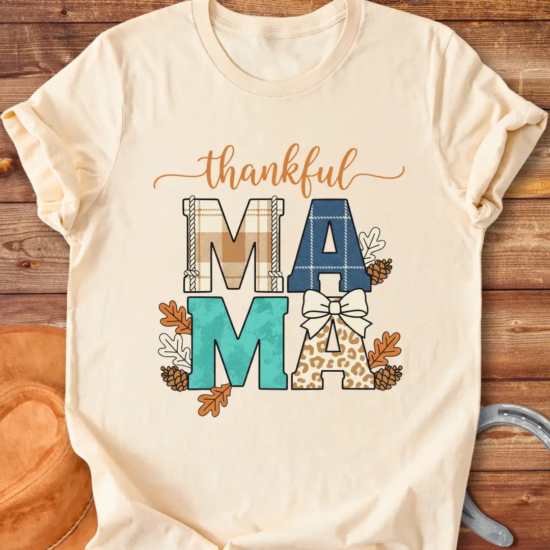 Patchwork Thankful Mama T-Shirt with autumn leaves, pinecones, and a bow on a black background. The design features the word 'thankful' and 'MAMA' in plaid and animal print letters.