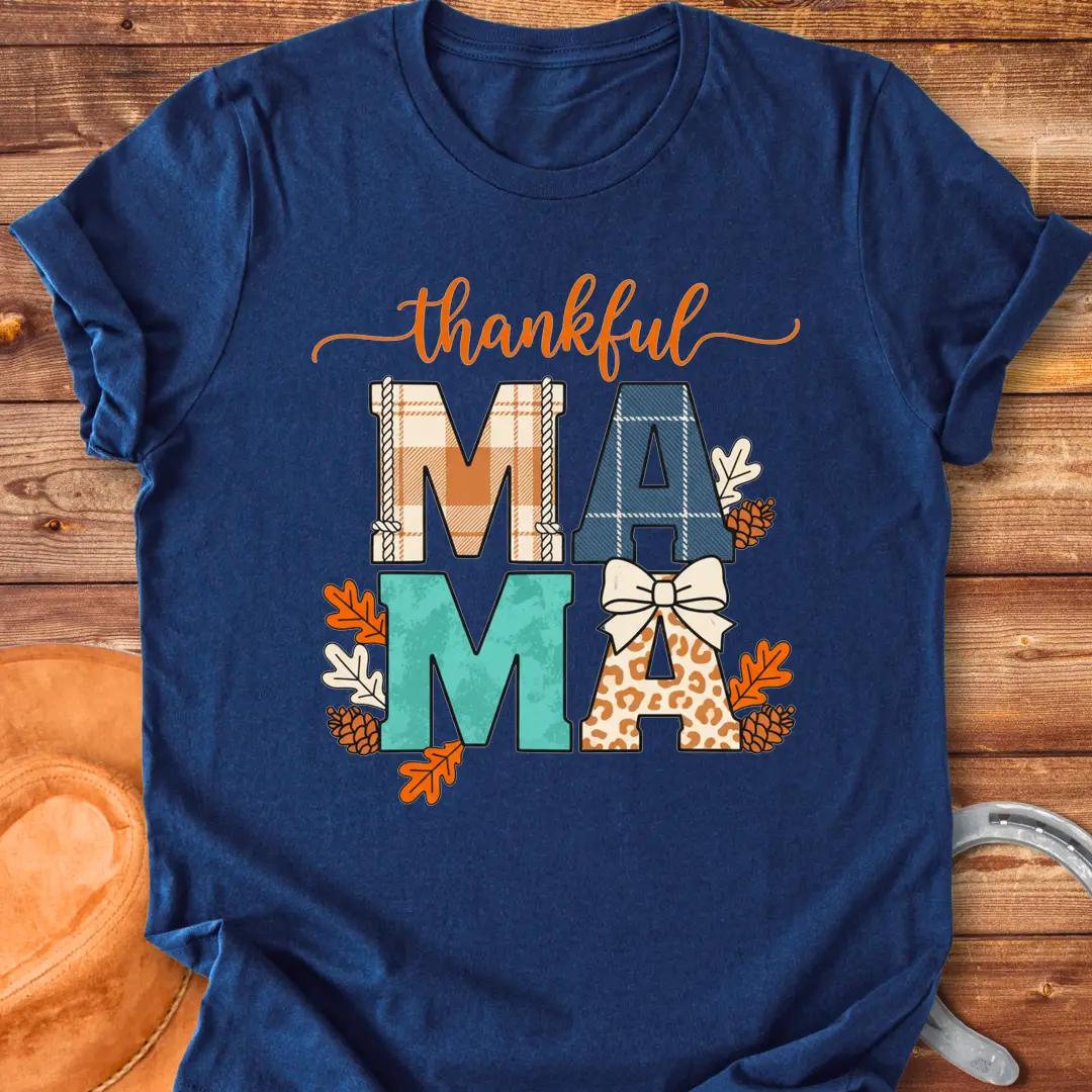 Patchwork Thankful Mama T-Shirt with autumn leaves, pinecones, and a bow on a black background. The design features the word 'thankful' and 'MAMA' in plaid and animal print letters.