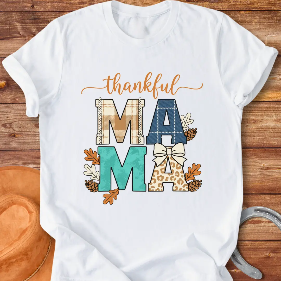 Patchwork Thankful Mama T-Shirt with autumn leaves, pinecones, and a bow on a black background. The design features the word 'thankful' and 'MAMA' in plaid and animal print letters.
