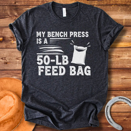 A graphic design on a t-shirt showing a humorous depiction of a feed bag labeled as a '50-lb' weight for a bench press.