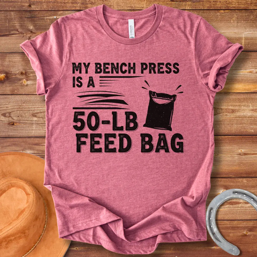 A graphic design on a t-shirt showing a humorous depiction of a feed bag labeled as a '50-lb' weight for a bench press.