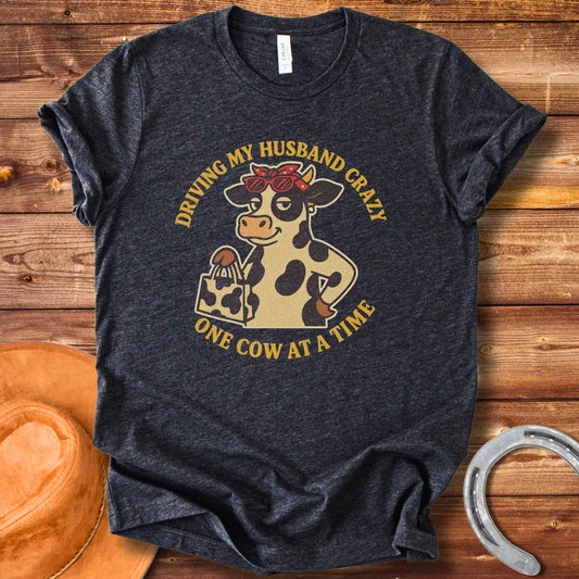A cartoon cow wearing sunglasses and a bandana, holding a shopping bag, with the text 'Driving My Husband Crazy One Cow At A Time' around it.