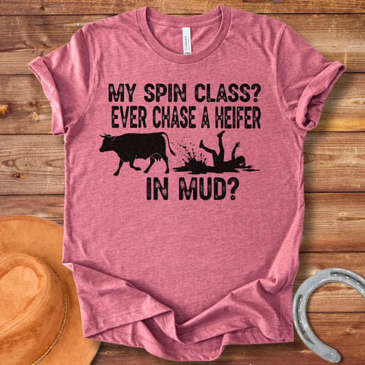 A funny illustration of a cow and a person falling in mud, with the text 'My spin class? Ever chase a heifer in mud?'