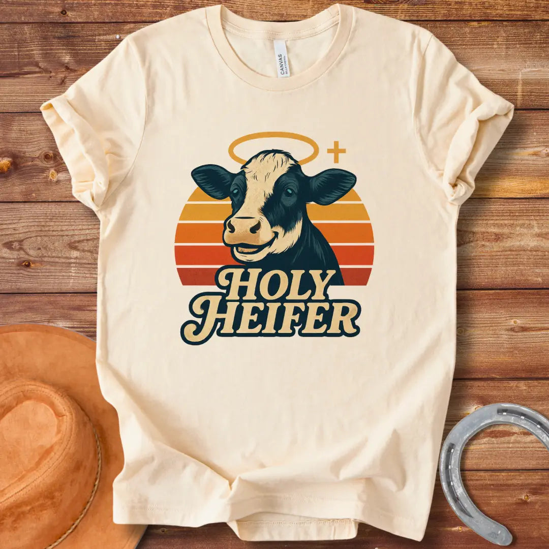 Holy Heifer T-Shirt from Cow Lovers Clothing, featuring a smiling cow with a halo and the text 'Holy Heifer' with a sunset background.