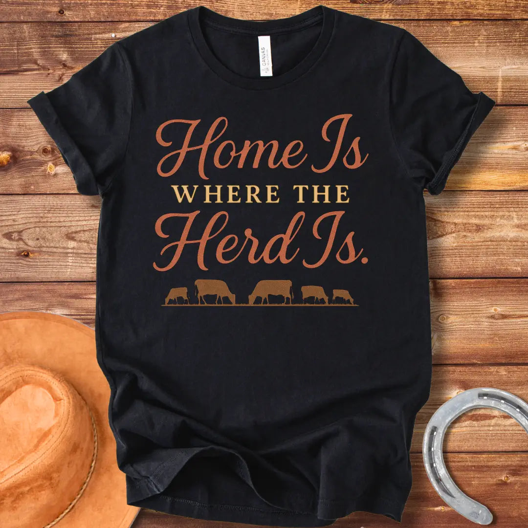 Orange text on a black t-shirt that says, "Home Is Where The Herd Is." Below the text is a silhouette of a herd of cows grazing.