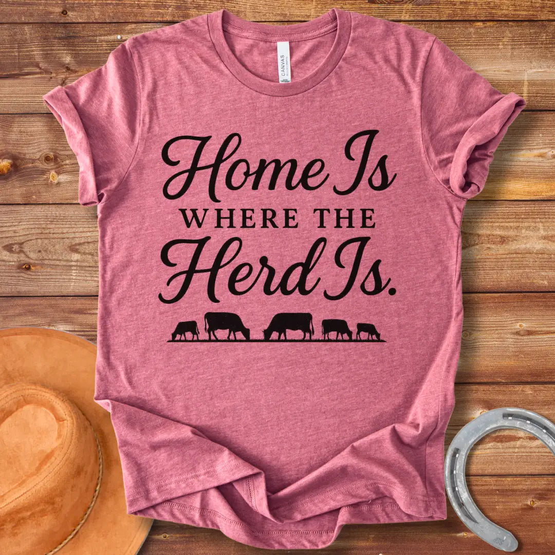 Orange text on a black t-shirt that says, "Home Is Where The Herd Is." Below the text is a silhouette of a herd of cows grazing.