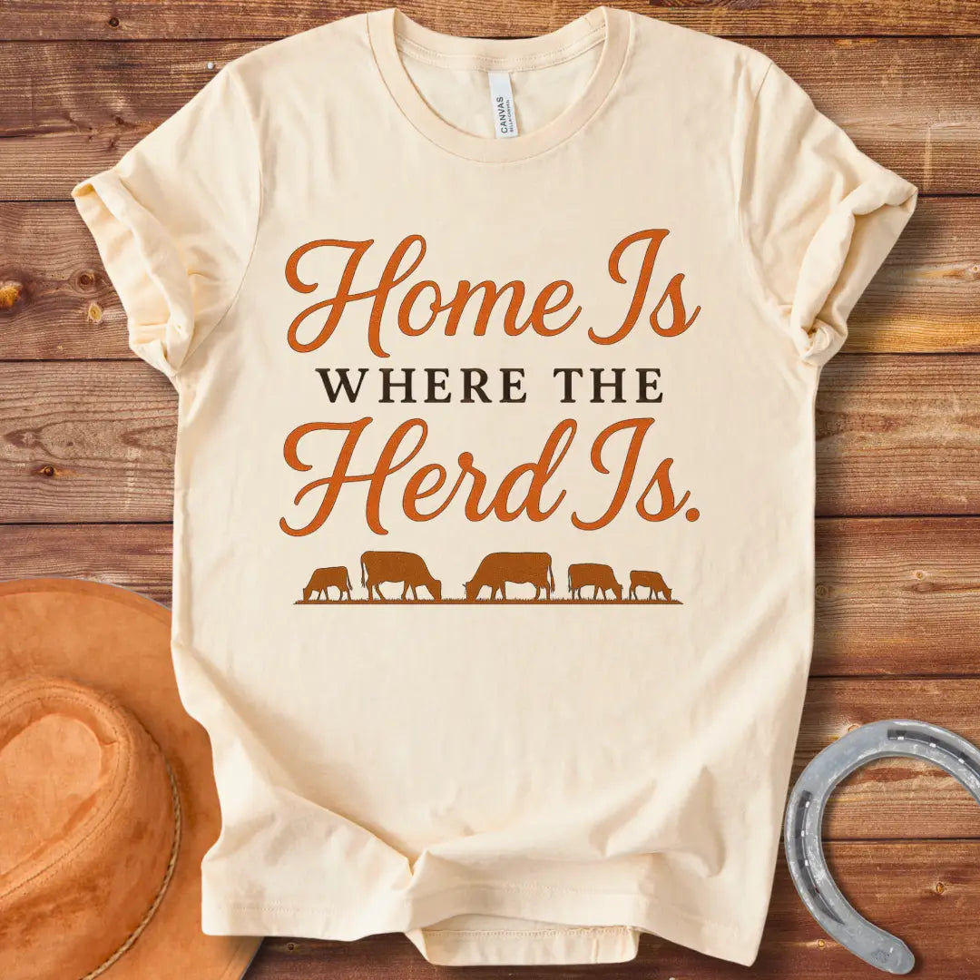 Orange text on a black t-shirt that says, "Home Is Where The Herd Is." Below the text is a silhouette of a herd of cows grazing.