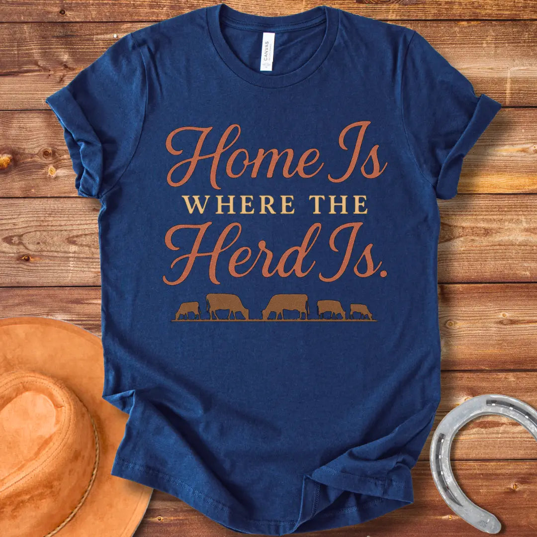 Orange text on a black t-shirt that says, "Home Is Where The Herd Is." Below the text is a silhouette of a herd of cows grazing.