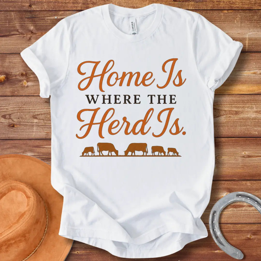 Orange text on a black t-shirt that says, "Home Is Where The Herd Is." Below the text is a silhouette of a herd of cows grazing.