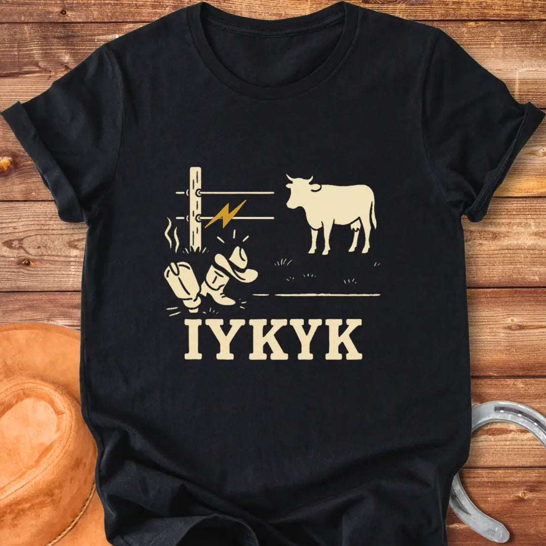 Graphic design on a t-shirt depicts a shocked cowboy hat and boots with a hot wire and a cow in the background, with the words IYKYK at the bottom