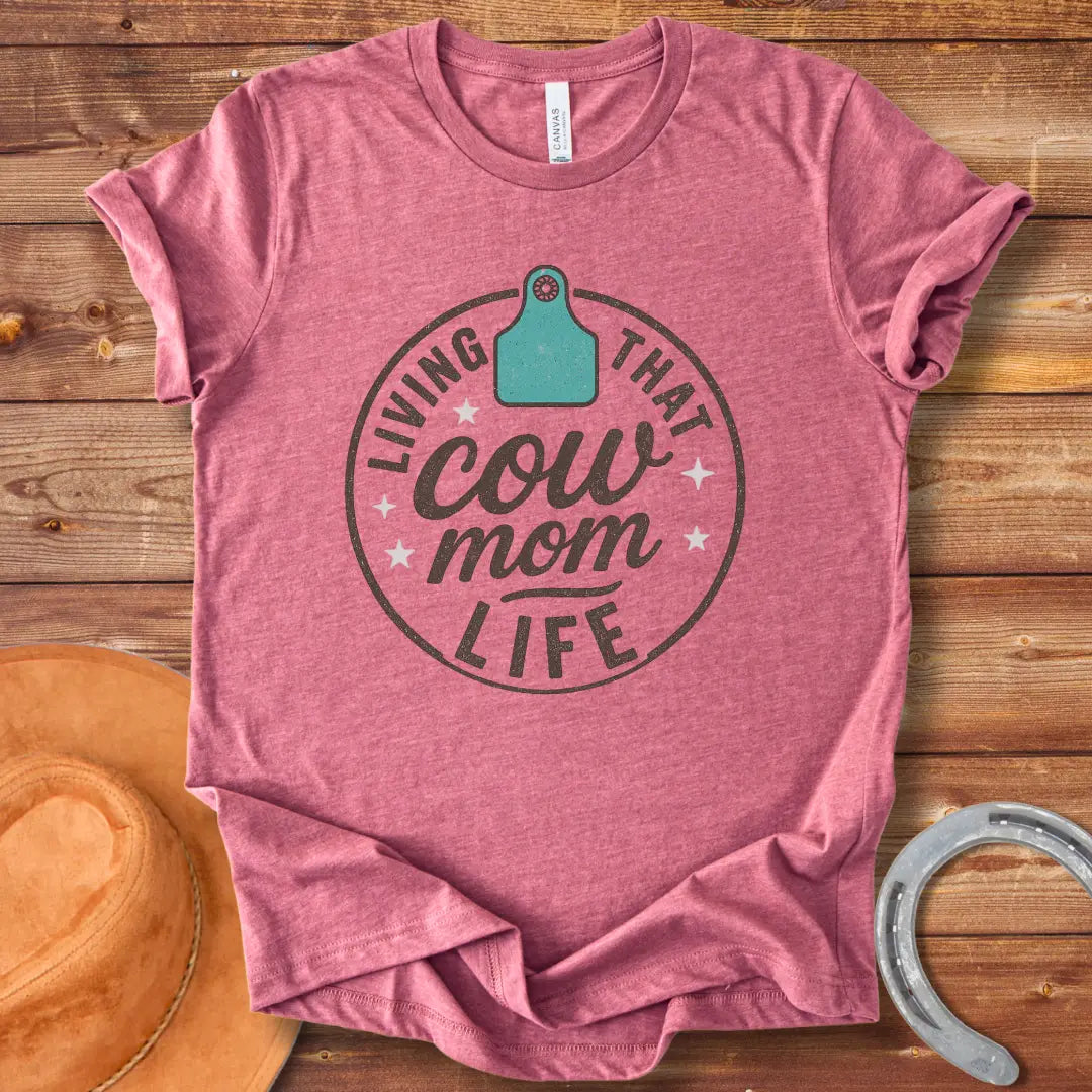 A round design featuring the text 'Living That Cow Mom Life' surrounding a cow tag graphic with orange stars.