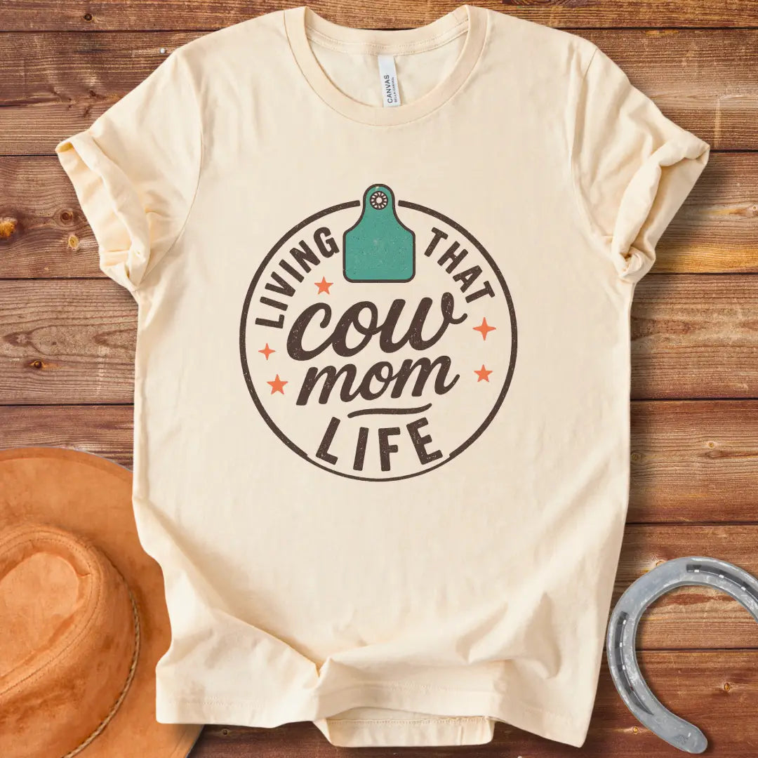 A round design featuring the text 'Living That Cow Mom Life' surrounding a cow tag graphic with orange stars.