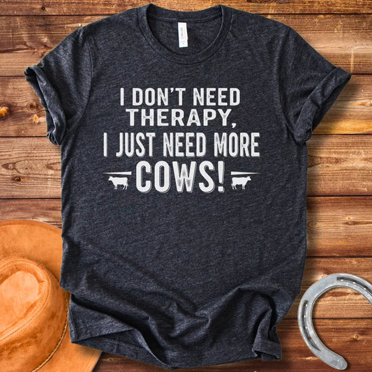 A black and white graphic tee design that says 'I DON'T NEED THERAPY, I JUST NEED MORE COWS!' with two small cow illustrations flanking the text.