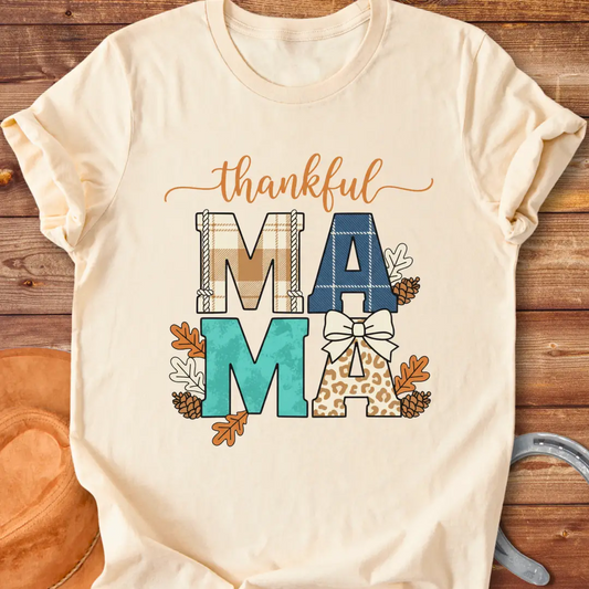 Patchwork Thankful Mama T-Shirt with autumn leaves, pinecones, and a bow on a black background. The design features the word 'thankful' and 'MAMA' in plaid and animal print letters.