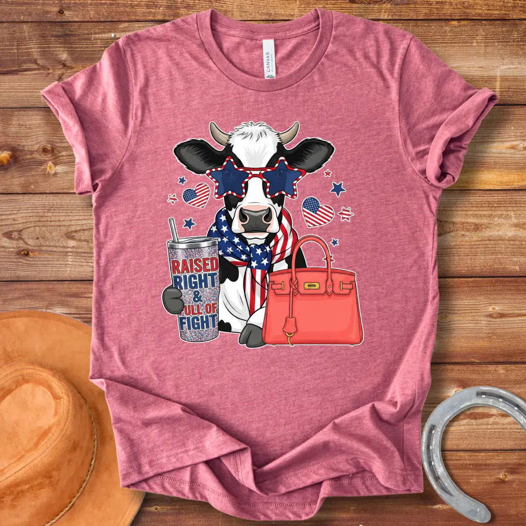 Patriotic cow t-shirt. Cow wearing star-shaped sunglasses, American flag scarf, and holding a tumbler that says 'Raised Right & Full of Fight', with a red purse.