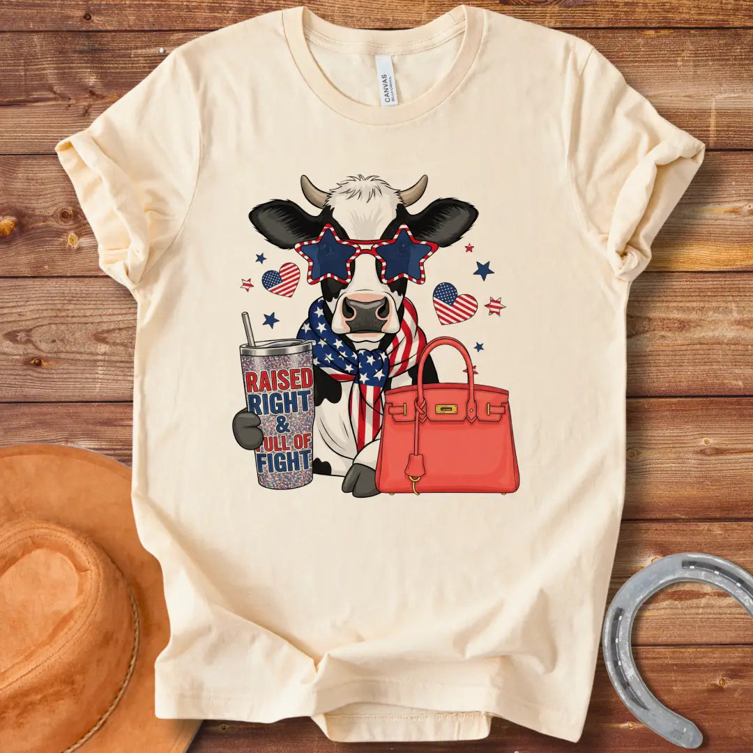 Patriotic cow t-shirt. Cow wearing star-shaped sunglasses, American flag scarf, and holding a tumbler that says 'Raised Right & Full of Fight', with a red purse.