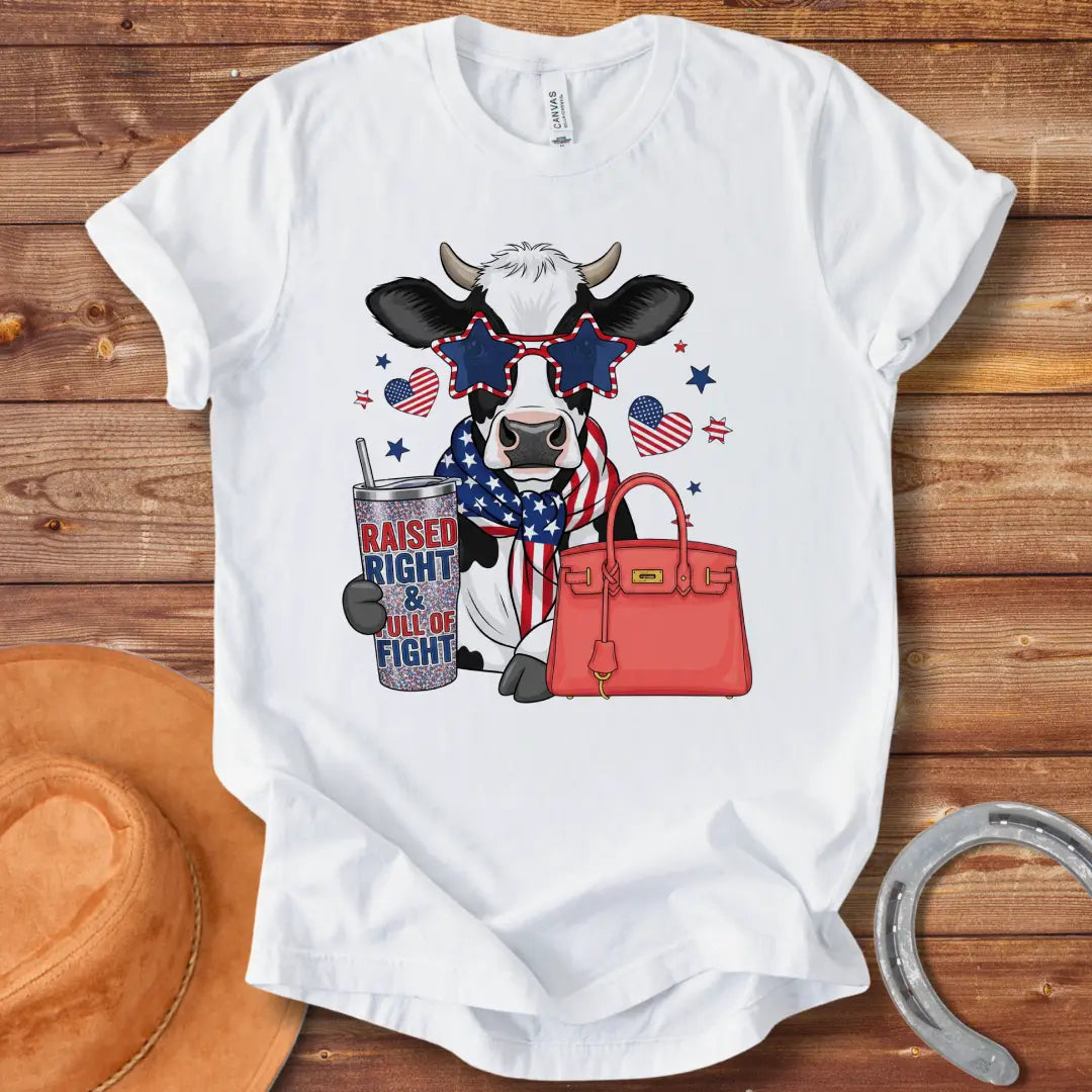 Patriotic cow t-shirt. Cow wearing star-shaped sunglasses, American flag scarf, and holding a tumbler that says 'Raised Right & Full of Fight', with a red purse.